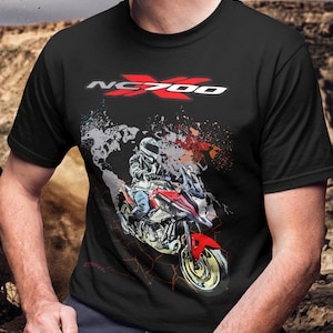 May include: Black t-shirt featuring a motorcycle graphic with the text "NC700" in red and white. The graphic depicts a rider on a red and white motorcycle with a splash effect. The shirt is a crew neck.
