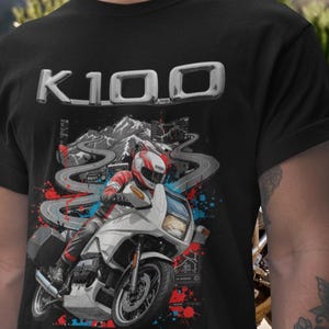 Pode incluir: Camiseta preta com um gráfico de motocicleta. O design apresenta uma motocicleta branca com um piloto, uma estrada sinuosa e o texto "K100". O gráfico tem detalhes em vermelho e azul e uma cordilheira ao fundo.