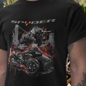 Könnte beinhalten: Schwarzes T-Shirt mit einer Grafik eines schwarzen dreirädrigen Motorrads, eines Bullen und einer Skyline. Das Wort "SPYDER" ist grau über der Grafik aufgedruckt. Rote Farbspritzer akzentuieren das Design.