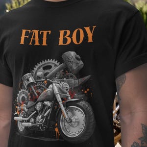 Op de afbeelding: Zwart t-shirt met de oranje tekst "FAT BOY" boven een motorfietsafbeelding. Het motorfietsontwerp bevat tandwielen, een hamer en oranje verfspatten. Het shirt is gemaakt van een zacht materiaal.