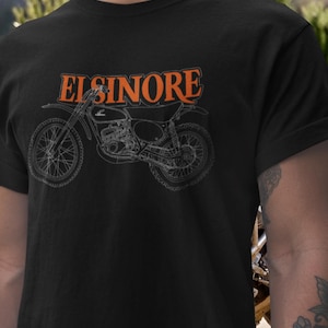 May include: Black t-shirt featuring a motorcycle graphic with the word "ELSINORE" in orange block letters above the motorcycle. The motorcycle is a detailed line drawing in gray. The shirt is a classic crew neck.