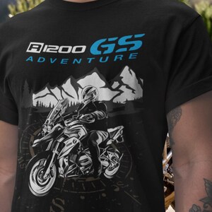 Pode incluir: Camiseta preta com um gráfico de motocicleta e o texto "R1200 GS Adventure" em azul e branco. O design inclui um motociclista em uma moto, montanhas e uma bússola.