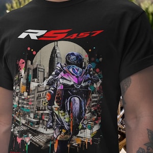 May include: Black t-shirt featuring a motorcycle graphic with a rider in a city setting. The design includes the text "RS457" in white and red. The artwork has colorful accents and a cityscape backdrop.