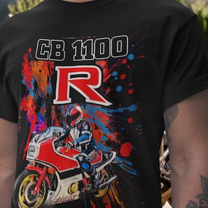 Pode incluir: T-shirt preto com um gráfico de motocicleta. O design apresenta o texto "CB 1100 R", uma moto vermelha e branca e um piloto, em um fundo colorido de respingos.