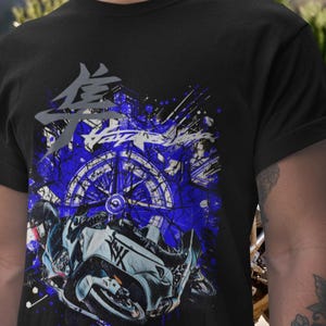 May include: Black t-shirt featuring a motorcycle graphic with blue and white accents. The design includes a stylized image of a motorcycle, a compass-like element, and Japanese characters. The overall theme is motorcycle apparel.