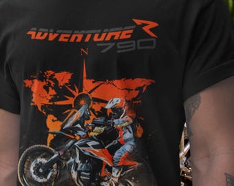 Puede incluir: Una camiseta negra con un gráfico naranja de un motociclista en movimiento sobre la silueta de un mapa mundial. El texto ADVENTURE R N 790 está impreso en naranja y gris sobre el gráfico, que incluye un elemento de diseño de rosa de los vientos.