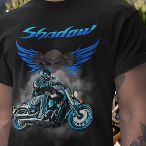 Könnte beinhalten: Schwarzes T-Shirt mit einem Motorrad-Motiv. Das Design zeigt einen Fahrer auf einem Motorrad, einen Totenkopf, Flügel und das Wort "Shadow" in Blau. Das Motorrad und die Akzente sind ebenfalls in Blautönen gehalten.