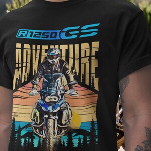 Peut inclure: T-shirt noir avec un motif de moto et le texte "R1250 GS ADVENTURE". Le design représente un motard sur une moto devant un coucher de soleil avec des arbres. Le texte est en bleu et or.