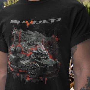 May include: Black t-shirt featuring a graphic of a Spyder motorcycle with a winding road and mountain backdrop. The design incorporates red and gray accents, with the word "SPYDER" across the top.