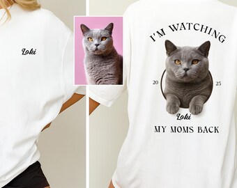 May include: White oversized t-shirt with the name "Loki" embroidered on the front. The back features a graphic of a gray cat with the text "I'M WATCHING" above and "Loki MY MOMS BACK" below the cat image.