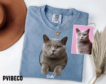 May include: A light blue Comfort Colors t-shirt featuring a gray cat illustration with the name "Loki" below. A small photo of the cat is next to the shirt. A gold necklace is draped on the shirt.