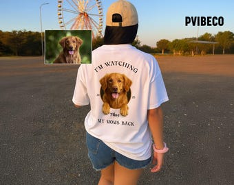 May include: White t-shirt with a golden retriever graphic. The shirt features the text "I'M WATCHING" above the dog image, with "Thor" and "MY MOMS BACK" below. A small image of the dog is also on the shirt. The person is wearing a tan baseball cap and denim shorts.