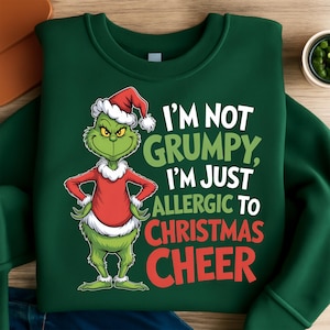Puede incluir: Sudadera verde oscuro con el personaje de Grinch con gorro de Papá Noel y atuendo rojo. El texto dice "I'M NOT GRUMPY, I'M JUST ALLERGIC TO CHRISTMAS CHEER". Una prenda de vestir con temática navideña.