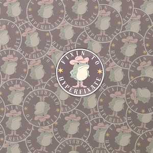 May include: A sticker with a cartoon frog in a cowboy hat and boots, with the text "FIXIN TO HYPERFIXATE" in a circular design. The background is a repeating pattern of the same design on a muted purple background.
