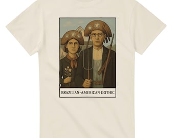 May include: Cream-colored t-shirt featuring a framed illustration of two people in hats, holding tools, and standing in front of a house. The text "BRAZILIAN-AMERICAN GOTHIC" is printed below the image.