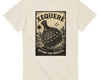 May include: Cream-colored t-shirt featuring a black and white illustration of a xeque, a Brazilian percussion instrument. The design includes the text "XEQUERE" above the instrument and "RITMO DO BRASIL" below it. A sun, cactus, and buildings are also in the design.