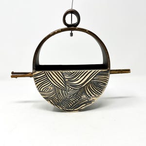 May include: A ceramic hanging planter with a black and white geometric pattern. The planter has a rounded shape and a metal frame with a hanging loop.