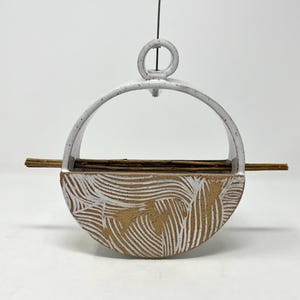 May include: A ceramic bird feeder with a white glaze and a textured, wave-like pattern. It features a circular handle and a wooden perch. The design combines functionality with artistic expression. The feeder is designed for outdoor use.