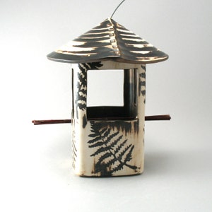 May include: A ceramic bird feeder with a pagoda-style roof and a rectangular body. The roof is black and white striped, and the body is cream-colored with black fern leaf designs. A small perch extends from the side.