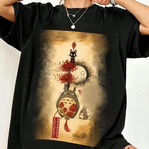 May include: Black t-shirt with a colourful illustration from the animated film "My Neighbour Totoro." The design includes Totoro, a black cat, a rabbit, and red flowers. The Japanese text "Totoro" is visible on a lantern.