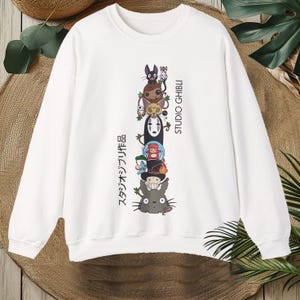 Puede incluir: Sudadera blanca con un dise&ntilde;o apilado de personajes de Studio Ghibli, incluyendo a Totoro y Sin Cara. El dise&ntilde;o incluye texto japon&eacute;s y las palabras "STUDIO GHIBLI" en negro. La sudadera tiene mangas largas y cuello redondo.