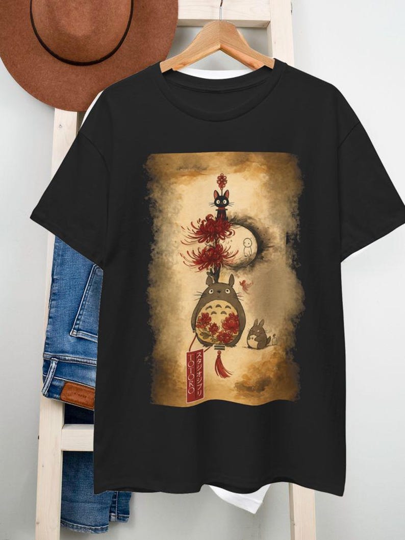 May include: Black t-shirt with a colourful illustration of animated film characters. The design includes a large, round creature with a red flower, a black cat, and a small rabbit. The shirt is hanging on a wooden rack.