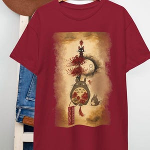 May include: Burgundy t-shirt with a vibrant illustration of characters from "My Neighbour Totoro." The design includes Totoro, a black cat, and other forest spirits. The shirt has a classic crew neck.