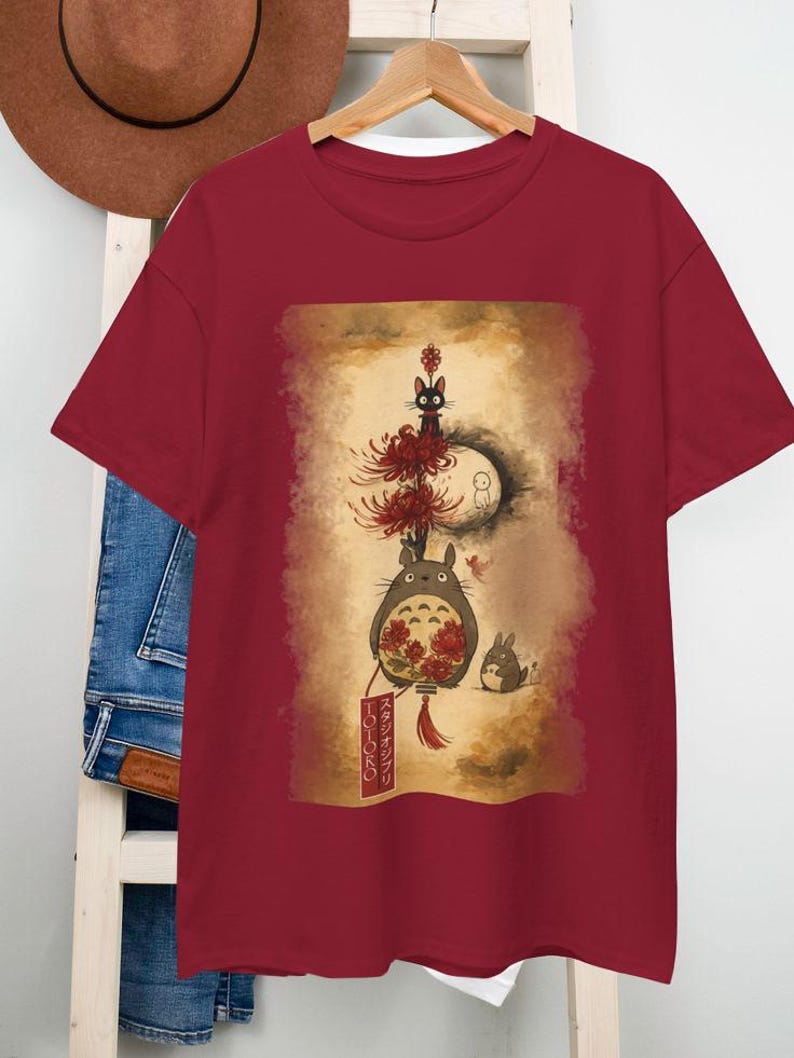 May include: Burgundy t-shirt with a vibrant illustration of characters from "My Neighbour Totoro." The design includes Totoro, a black cat, and other forest spirits. The shirt has a classic crew neck.
