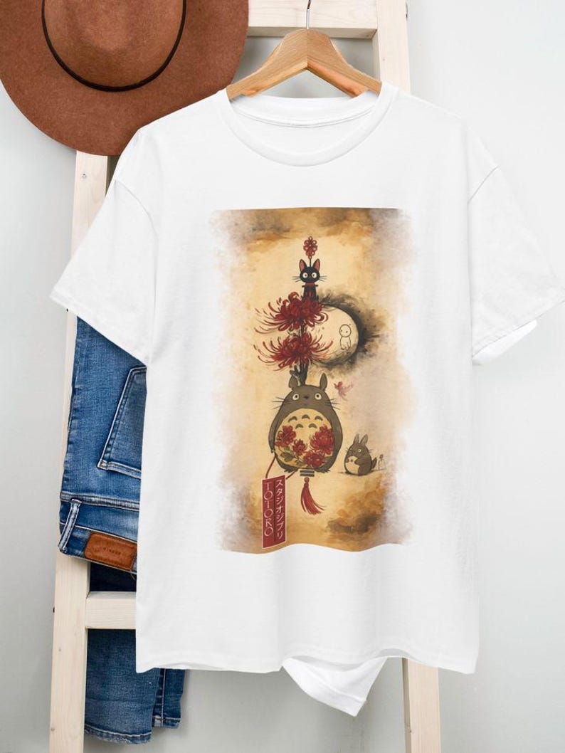 May include: White t-shirt with a Japanese-inspired illustration. The design features a large, round creature with red flowers, a black cat, a rabbit, and a moon. Japanese text is on a red banner. The artwork has a watercolour style.