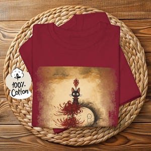 May include: A maroon t-shirt with a graphic featuring a black cat with red accents, surrounded by red flowers and a small white figure. The shirt is made of 100% cotton.