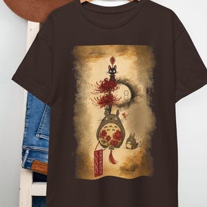 May include: A brown t-shirt with a graphic print of characters from the animated film, My Neighbour Totoro. The design features Totoro, a black cat, and other forest spirits, with Japanese text on a banner. The artwork is set against a sepia-toned background.