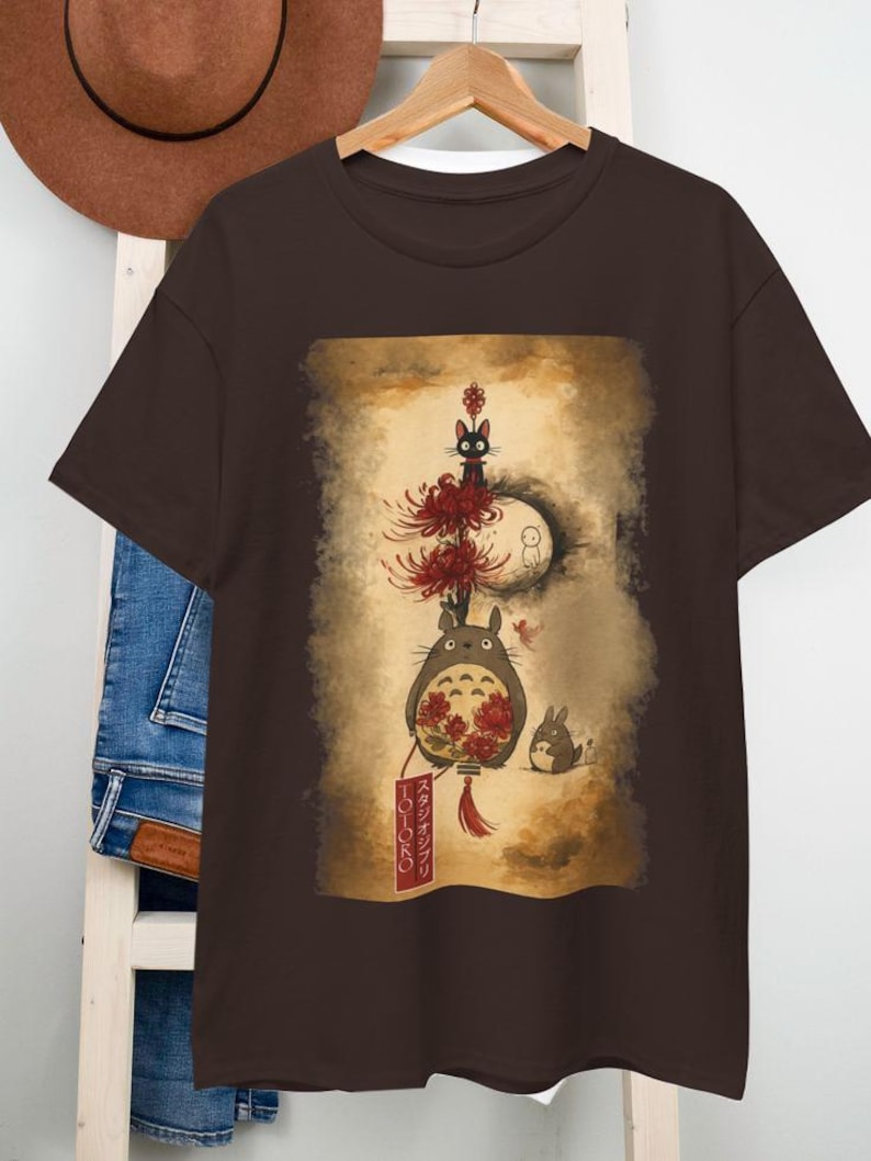 May include: A brown t-shirt with a graphic print of characters from the animated film, My Neighbour Totoro. The design features Totoro, a black cat, and other forest spirits, with Japanese text on a banner. The artwork is set against a sepia-toned background.