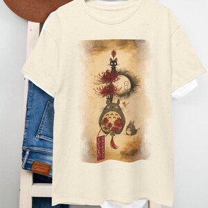May include: A cream-coloured t-shirt with a whimsical illustration. The design features a large, grey, furry creature with red flowers, a black cat, and a small rabbit. The artwork has a vintage, artistic style, with Japanese text on a red banner.