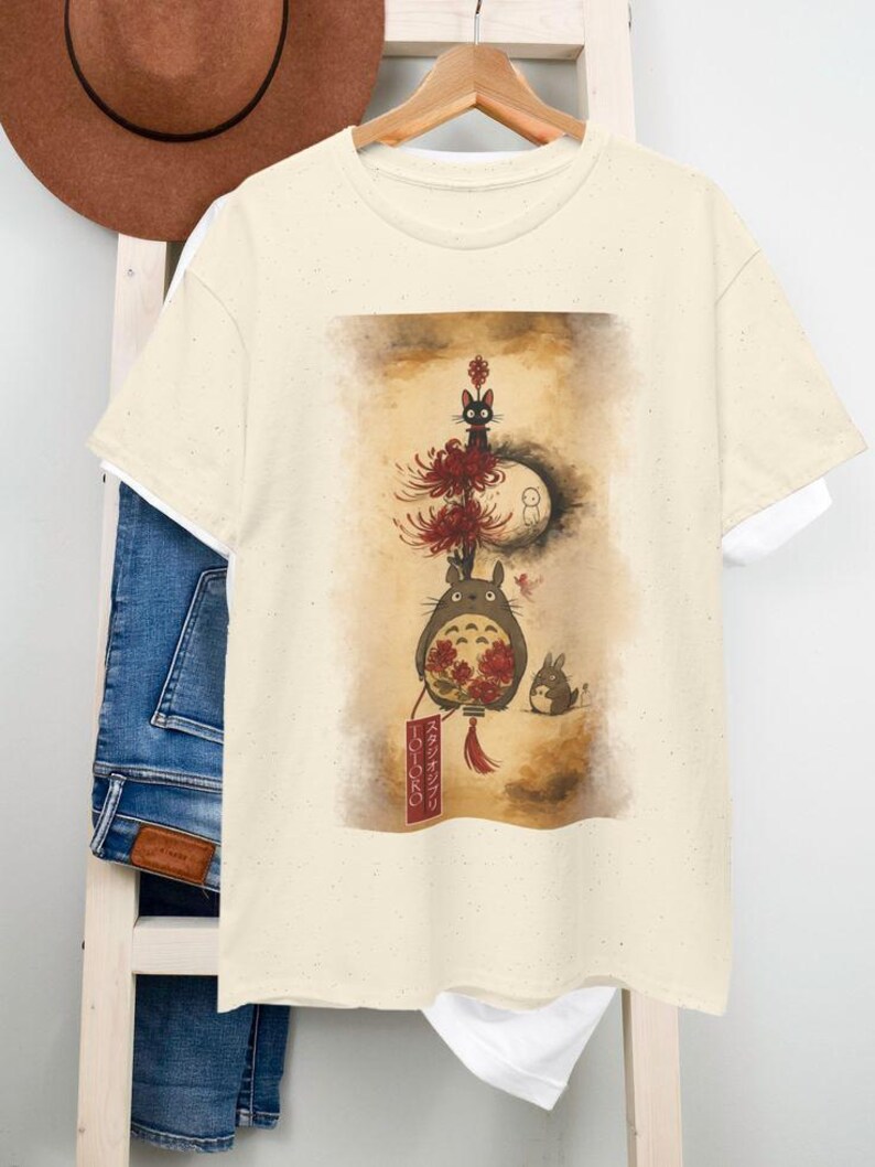 May include: A cream-coloured t-shirt with a whimsical illustration. The design features a large, grey, furry creature with red flowers, a black cat, and a small rabbit. The artwork has a vintage, artistic style, with Japanese text on a red banner.