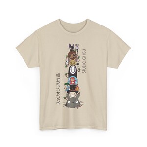 Puede incluir: Camiseta beige con una pila vertical de personajes de Studio Ghibli. El dise&ntilde;o incluye personajes como Totoro y Sin Rostro, con texto japon&eacute;s y "STUDIO GHIBLI" impreso verticalmente en el lado derecho.