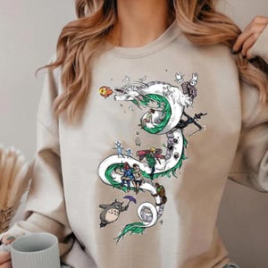 May include: Beige sweatshirt featuring a white dragon design with green accents and various cartoon characters. The dragon winds down the front of the shirt, with characters riding on its back and around its body. The design includes a Totoro character holding an umbrella.