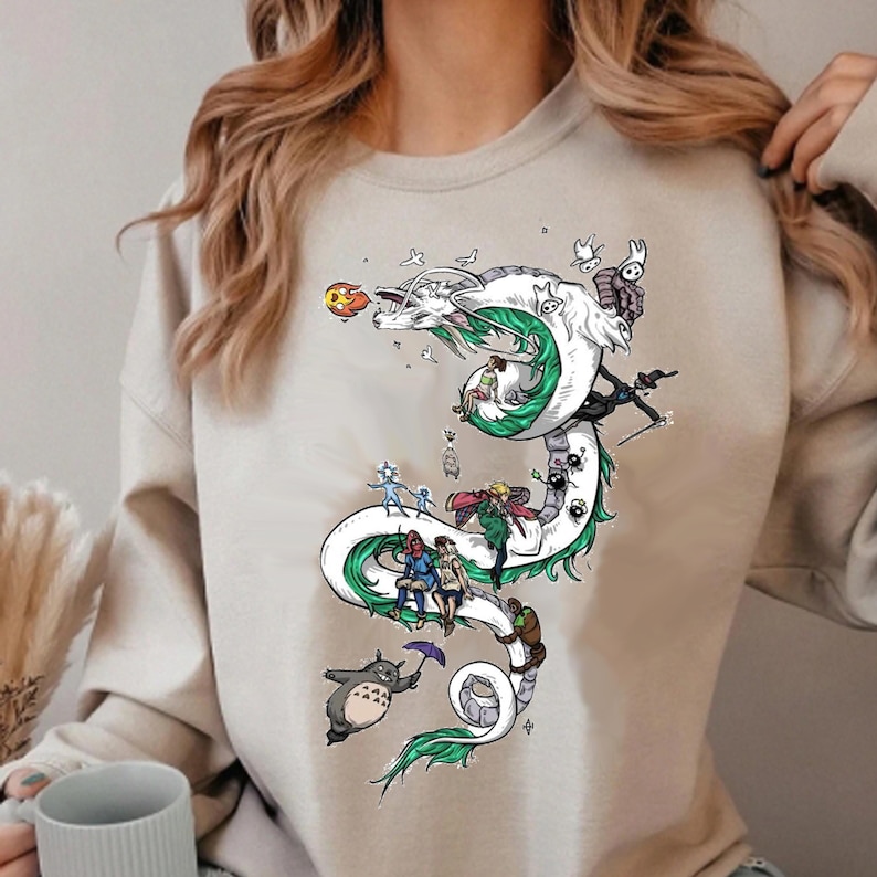May include: Beige sweatshirt featuring a white dragon design with green accents and various cartoon characters. The dragon winds down the front of the shirt, with characters riding on its back and around its body. The design includes a Totoro character holding an umbrella.
