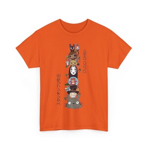Puede incluir: Camiseta naranja con un dise&ntilde;o apilado de dibujos animados de personajes de Studio Ghibli, incluyendo a Totoro y Sin Rostro. Texto japon&eacute;s impreso verticalmente a la izquierda, con "STUDIO GHIBLI" a la derecha. Un dise&ntilde;o vibrante y colorido en una camiseta cl&aacute;sica.