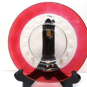 May include: A decorative plate with a vibrant red rim and a white center with a pattern of small, raised circles. A dark brown object is centered on the plate, and the plate is supported by a black stand.