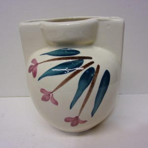 May include: Cream-colored ceramic vase with a rounded body and a small, rectangular top section. The vase is decorated with stylized teal leaves, brown stems, and pink flower accents. A small, shallow bowl is integrated into the top.