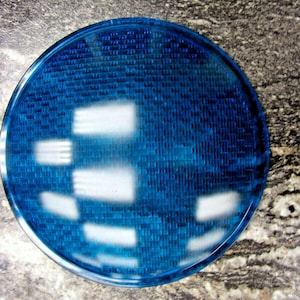 May include: A round, transparent blue object with a textured surface and a grid-like pattern. The object has a series of white rectangular shapes. The object is on a speckled gray and brown surface.