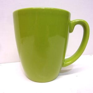 May include: A solid green ceramic mug with a curved handle. The mug has a glossy finish and a simple, modern design. The mug is a bright, vibrant shade of green.