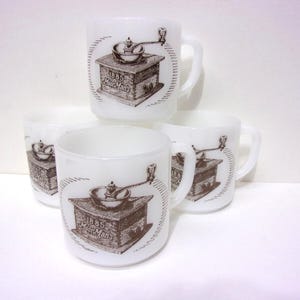 May include: Set of four white ceramic mugs. Each mug features a brown illustration of a coffee grinder with the text "1885 Hour Man's" in a circular design. The mugs have a classic handle design.