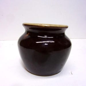 May include: A small, round, dark brown ceramic jar with a light tan rim. The jar has a glossy finish and a slightly flared opening. The jar is likely used for storage or decoration.