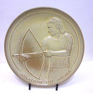 May include: A tan ceramic plate featuring a relief of an archer with a bow and arrow. The plate is round with the words "JONATHAN THE ARCHER" around the bottom edge. The plate is displayed on a black metal stand.