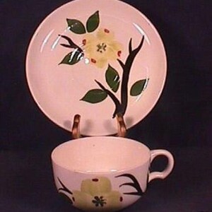 May include: A vintage teacup and saucer set. The set features a light pink background with a floral design in yellow, green, and black. The saucer is round, and the teacup has a handle. The set is displayed on a dark background.
