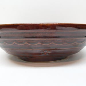 May include: A brown ceramic bowl with a glossy finish. The bowl has a wide rim and a series of horizontal ridges with a scalloped design. A small floral pattern is visible near the base. The bowl is likely used for serving food.