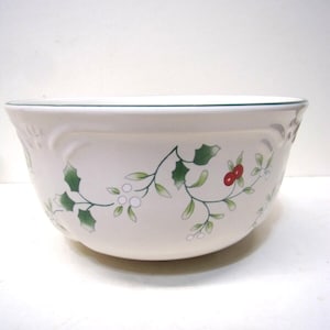 May include: A white ceramic bowl with a green rim and a decorative holly and berry pattern. The bowl has a curved shape and is suitable for serving or display. The design includes green leaves and red berries.