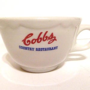 May include: White ceramic cup with a handle, featuring the red cursive text "Cobb's" above the blue text "Country Restaurant." The cup has a scalloped rim and is likely for serving beverages. The design suggests a diner or restaurant setting.