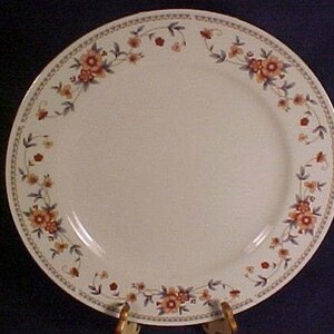 May include: A white ceramic dinner plate with a floral design. The plate features a border of small orange and red flowers with green leaves. The rim has a decorative pattern. The plate is on a gold stand.
